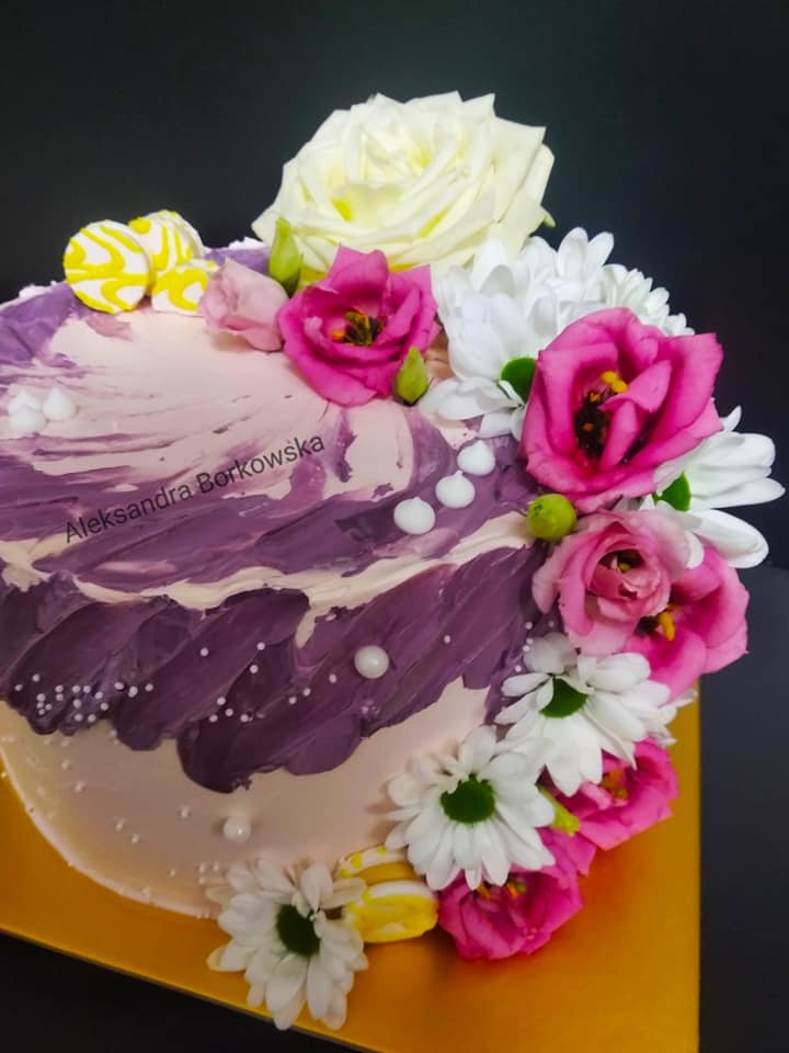 Dekorowany tort z fioletowo-białym kremem, ozdobiony świeżymi kwiatami: białą różą, różowymi eustomami i białymi margaretkami, oraz małymi, żółtymi makaronikami.