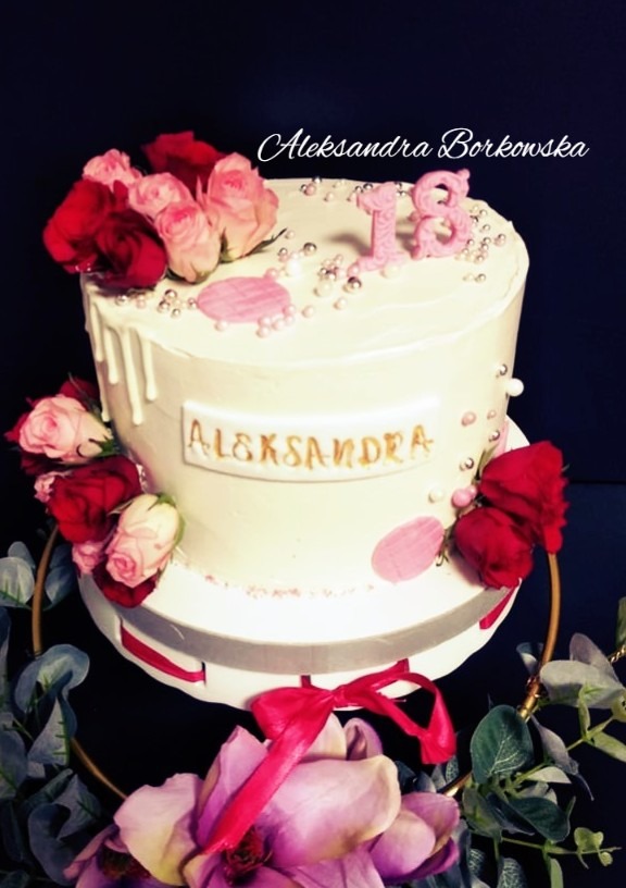 Dwupoziomowy tort urodzinowy z białym lukrem, zdobiony świeżymi różami w odcieniach różu i czerwieni, srebrnymi perełkami i napisem 'Aleksandra' w złotym kolorze, z topperem w kształcie liczby 18.