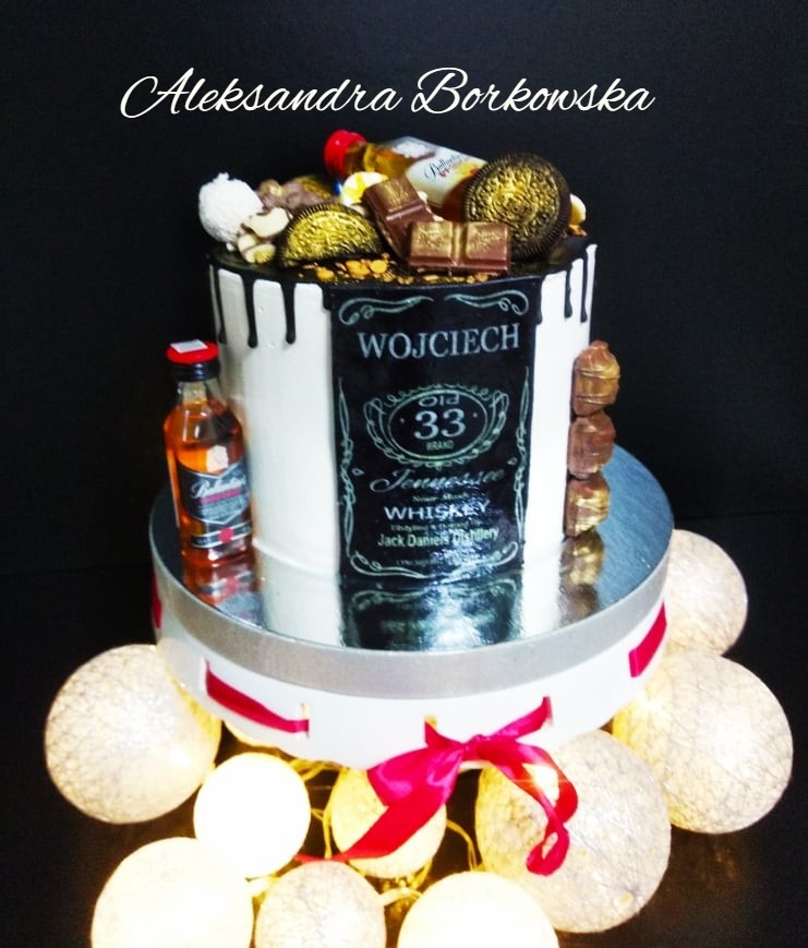 Dwupiętrowy tort urodzinowy w stylu whisky z buteleczką Ballantine's, czekoladkami i złotymi Oreo na wierzchu, dekorowany czarną polewą i personalizowaną etykietą 'Wojciech 33', otoczony...