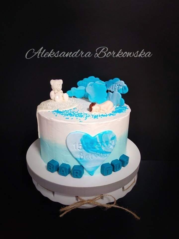 Tort na 15. urodziny z dekoracją w odcieniach bieli i błękitu: figurka śpiącego dziecka, pluszowy miś, chmurki i napis z kostek. Na torcie widnieje imię 'Aleksandra Borkowska'.