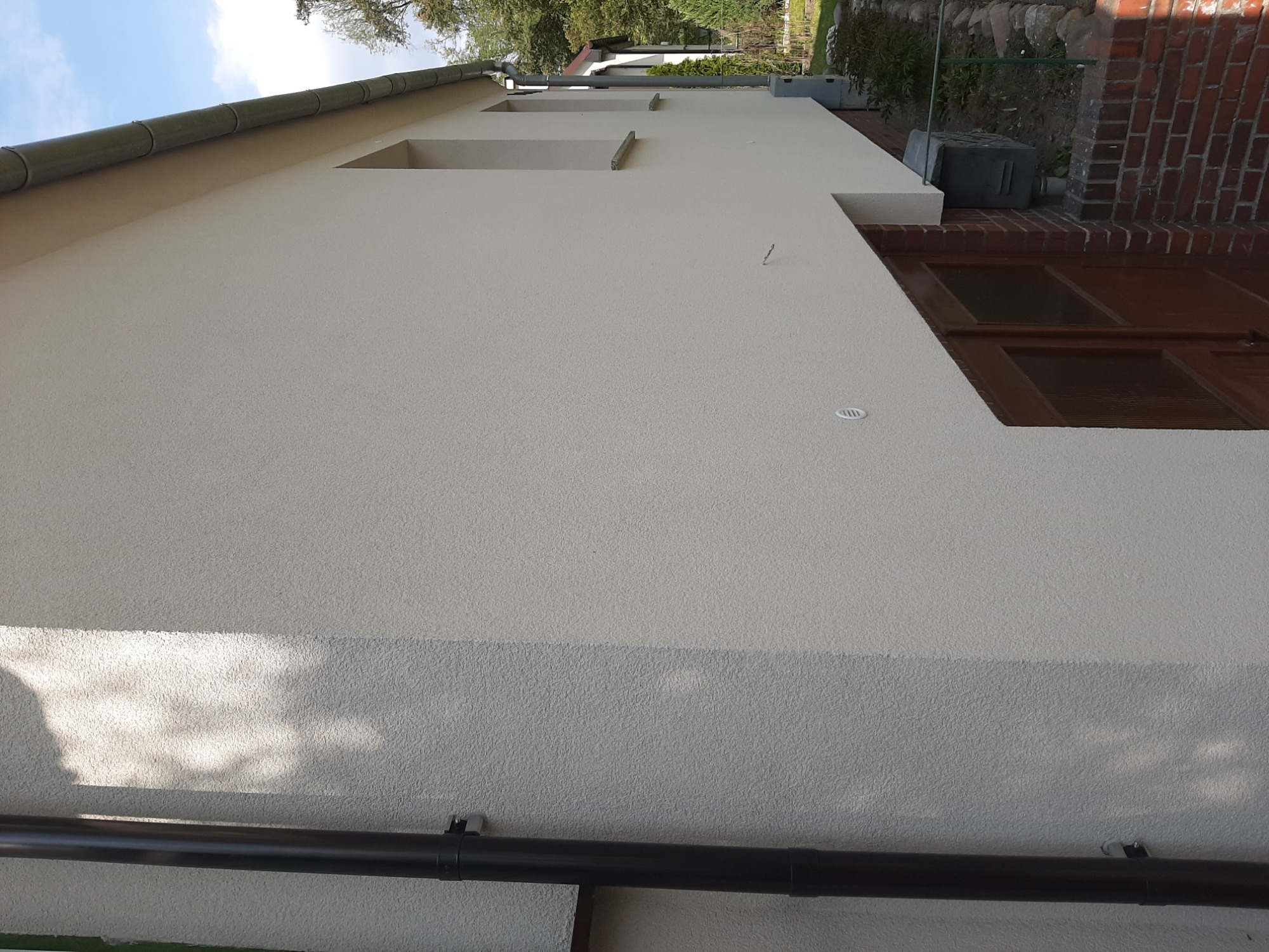 Świeżo otynkowana, jasna elewacja budynku jednorodzinnego z widocznymi elementami drenażu i fragmentem muru z czerwonej cegły.