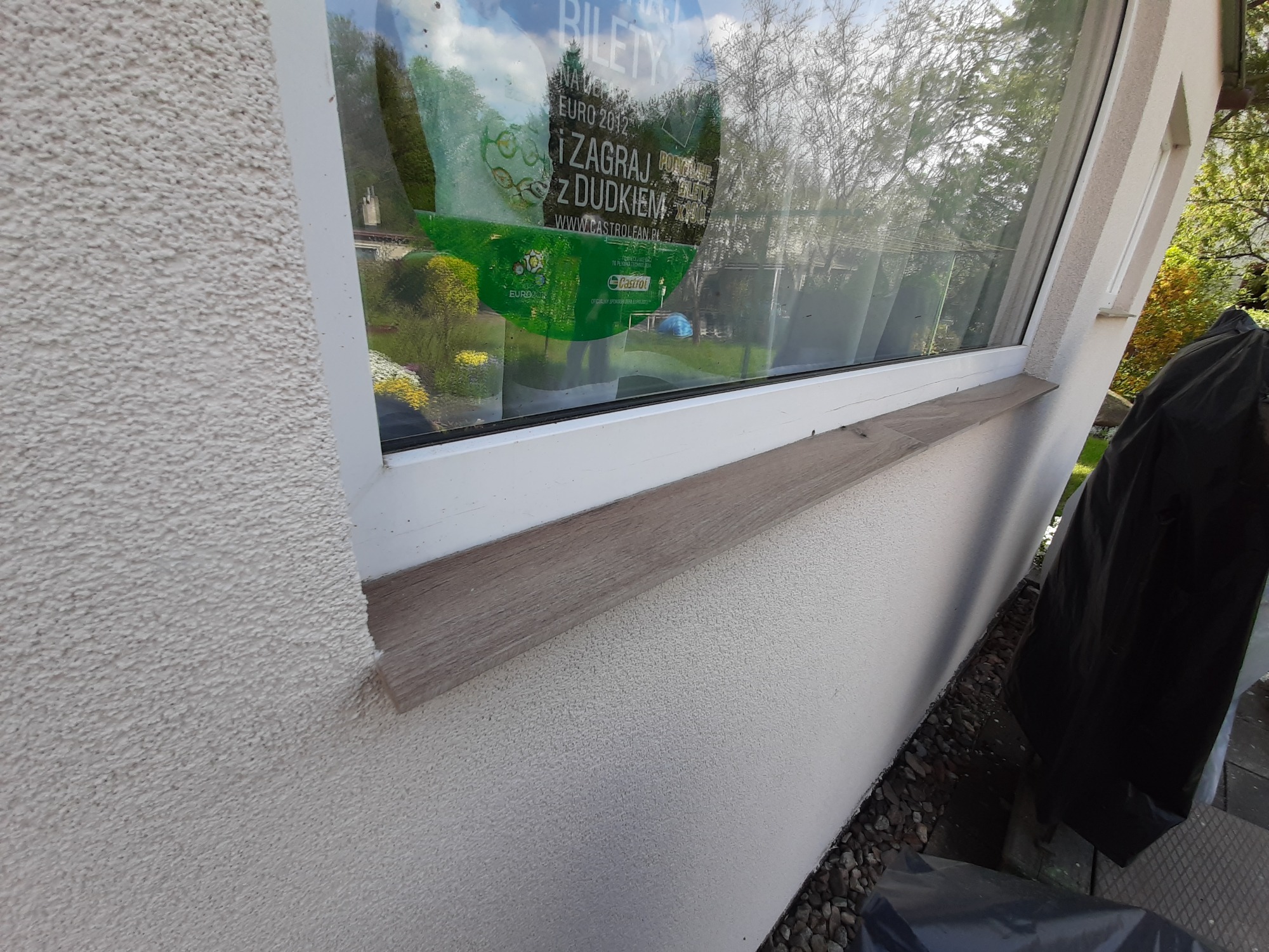 Fragment elewacji budynku z oknem, gdzie widać strukturę tynku baranka, białą ramę okienną z drewnianym parapetem i naklejkę na szybie okna.