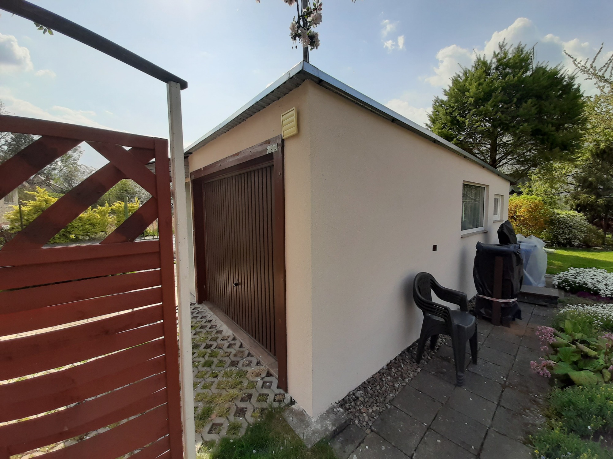 Otynkowany garaż z brązową bramą segmentową, widoczny zza drewnianego płotu, z krzesłem i roślinnością obok, na tle błękitnego nieba.