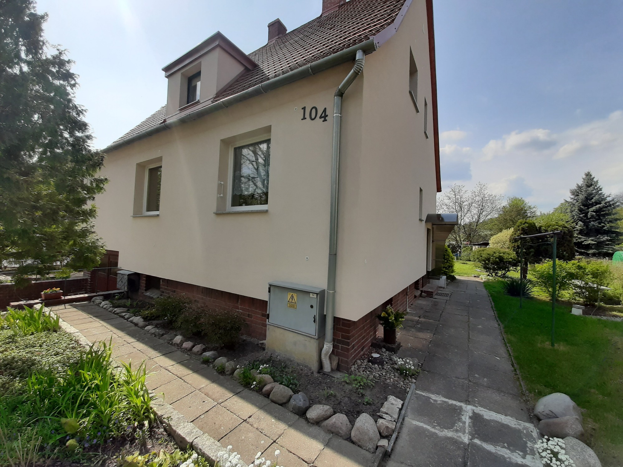 Dwupiętrowy dom z beżową elewacją, numerem 104 i brązowym dachem, widziany z boku, z chodnikiem z kamiennych płyt i rabatą kwiatową.