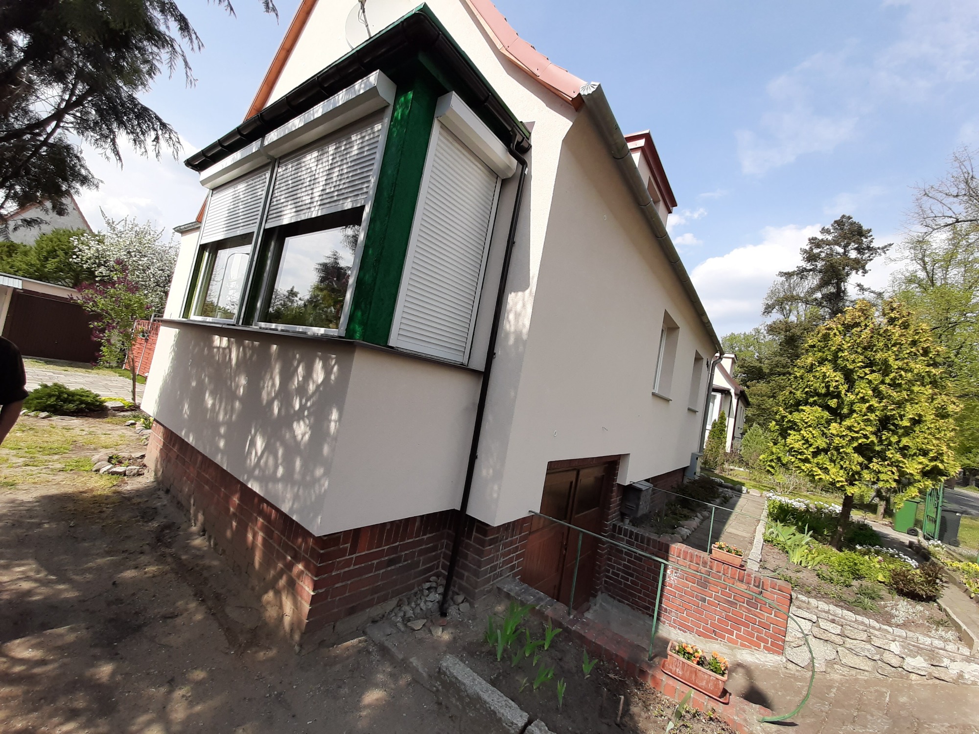 Jasna elewacja domu z ceglaną podmurówką i zielonymi elementami okiennymi, rolety zewnętrzne częściowo opuszczone, widok z boku na ogród z roślinami.