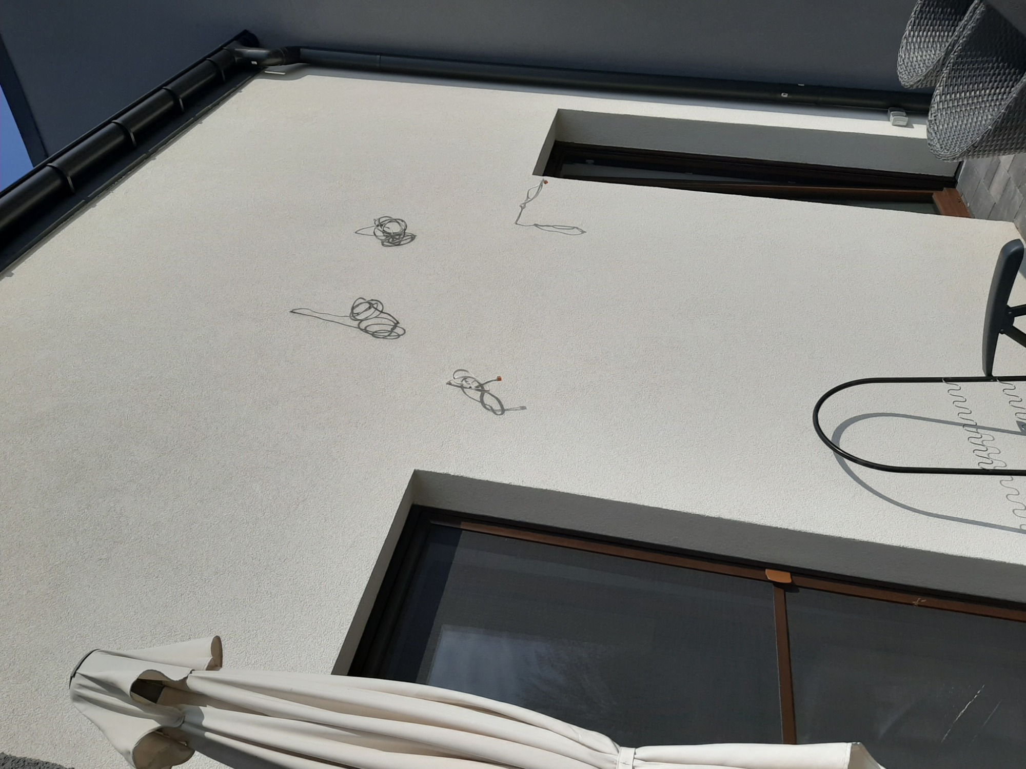 Fragment elewacji budynku z oknami i wystającymi przewodami elektrycznymi, widoczne elementy parasola i mebli balkonowych.