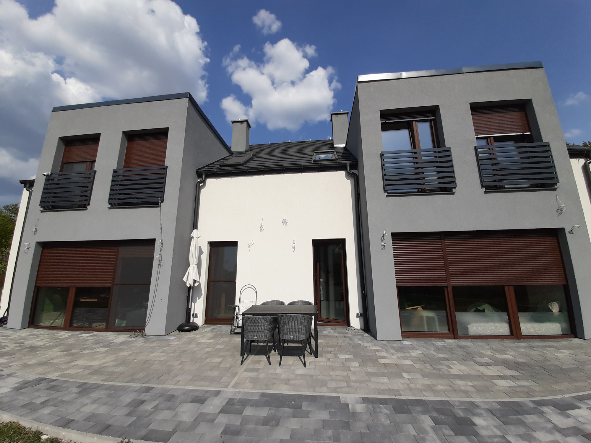 Nowoczesny dom z szarą elewacją, brązowymi roletami i szarym brukowanym podjazdem, widok na białą ścianę z ozdobami, stół i krzesła ogrodowe na pierwszym planie, błękitne niebo z chmurami w tle.