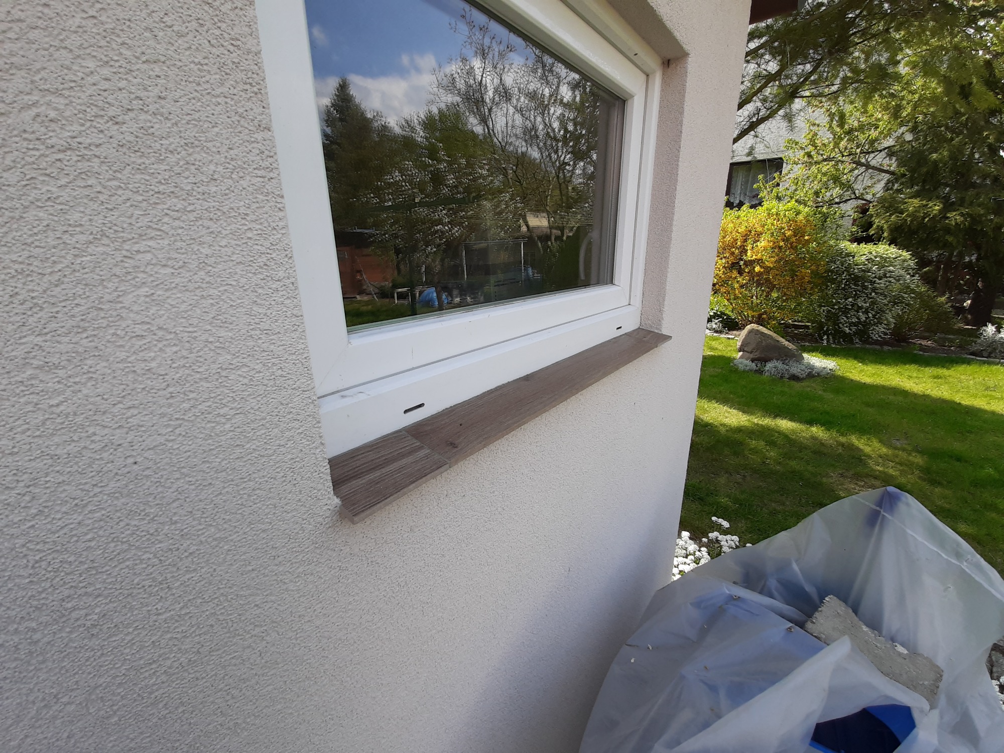 Fragment ściany budynku z oknem i drewnianym parapetem, widoczna faktura elewacji, w tle ogród z kwitnącymi krzewami, u dołu folia budowlana.