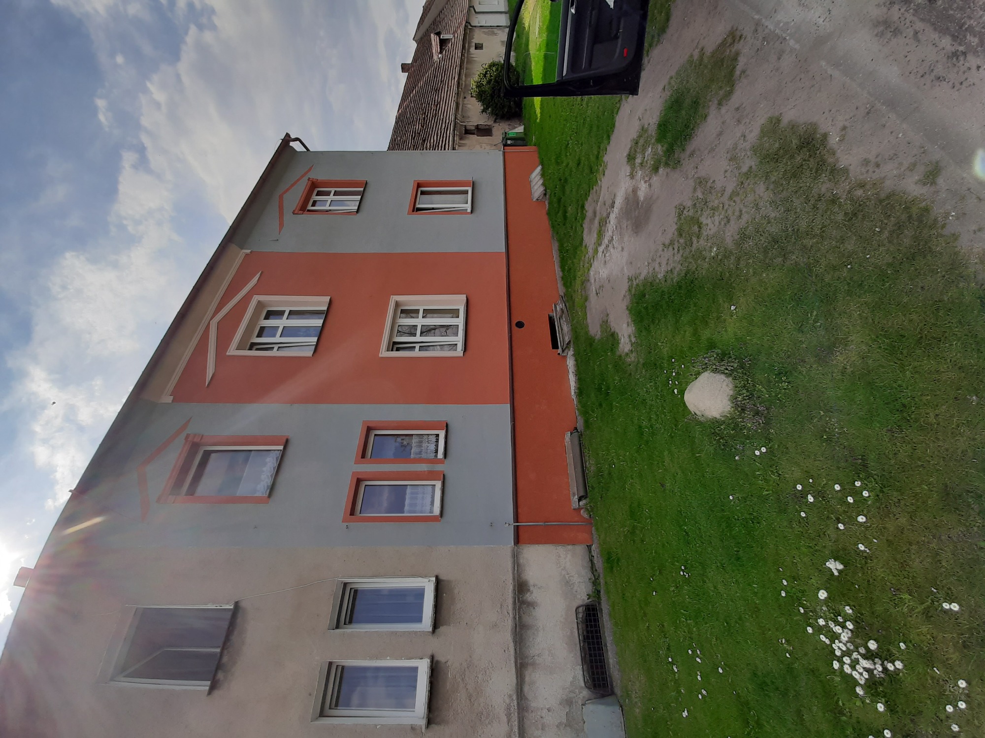 Dwukolorowa elewacja domu z widocznymi oknami, trawnikiem i fragmentem otwartych drzwi samochodu.