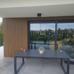 Nowoczesny taras z szarych płyt, drewnianą ścianą z pionowych paneli i dużymi przesuwnymi drzwiami szklanymi, stół ogrodowy z dekoracją roślinną.