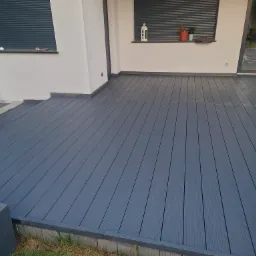 Nowoczesny taras kompozytowy w odcieniach szarości, z widocznymi ryflowanymi deskami, donicą z białymi kamieniami i fragmentem elewacji z oknami.