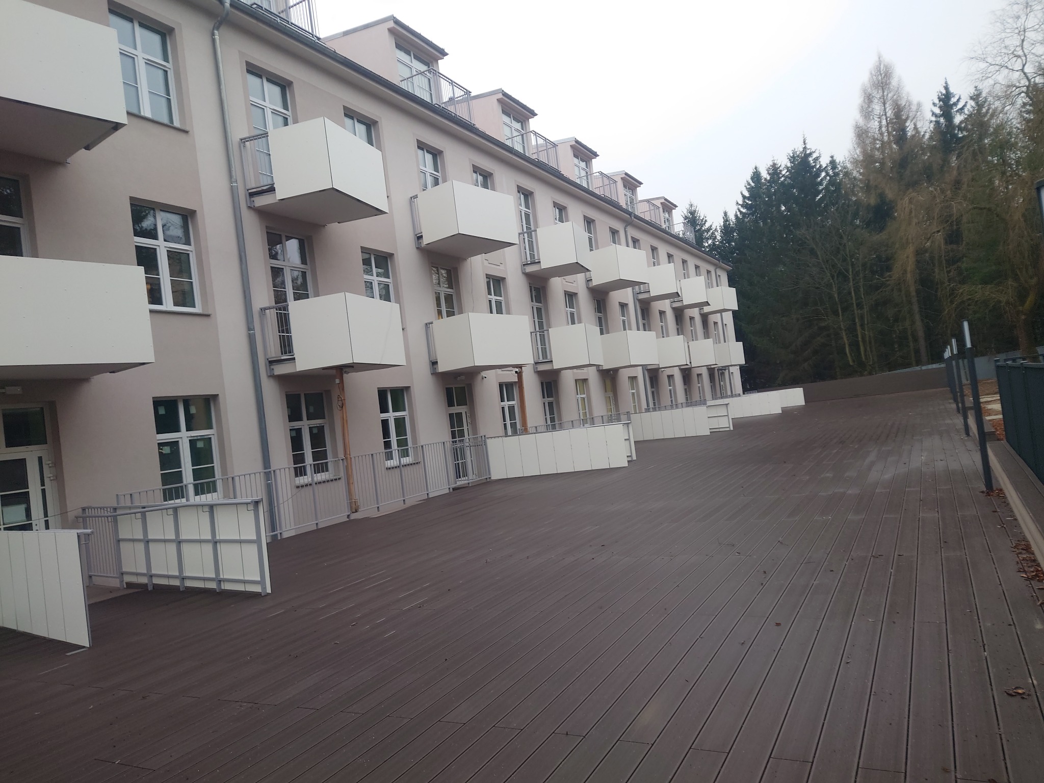 Rozległy taras kompozytowy przed budynkiem apartamentowym z białymi balkonami, widok z perspektywy, pochmurny dzień.