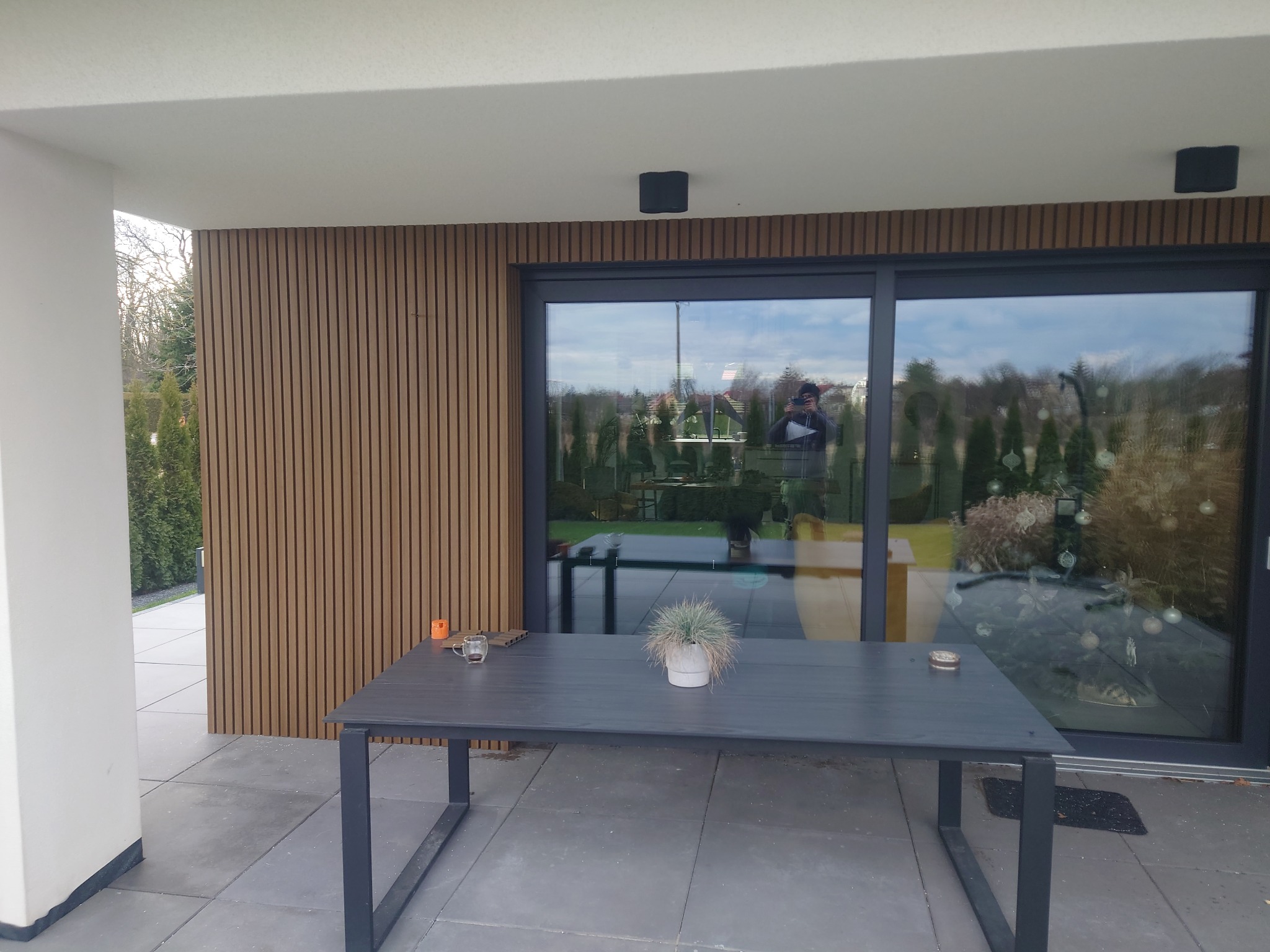 Nowoczesny taras z szarych płyt, drewnianą ścianą z pionowych paneli i dużymi przesuwnymi drzwiami szklanymi, stół ogrodowy z dekoracją roślinną.