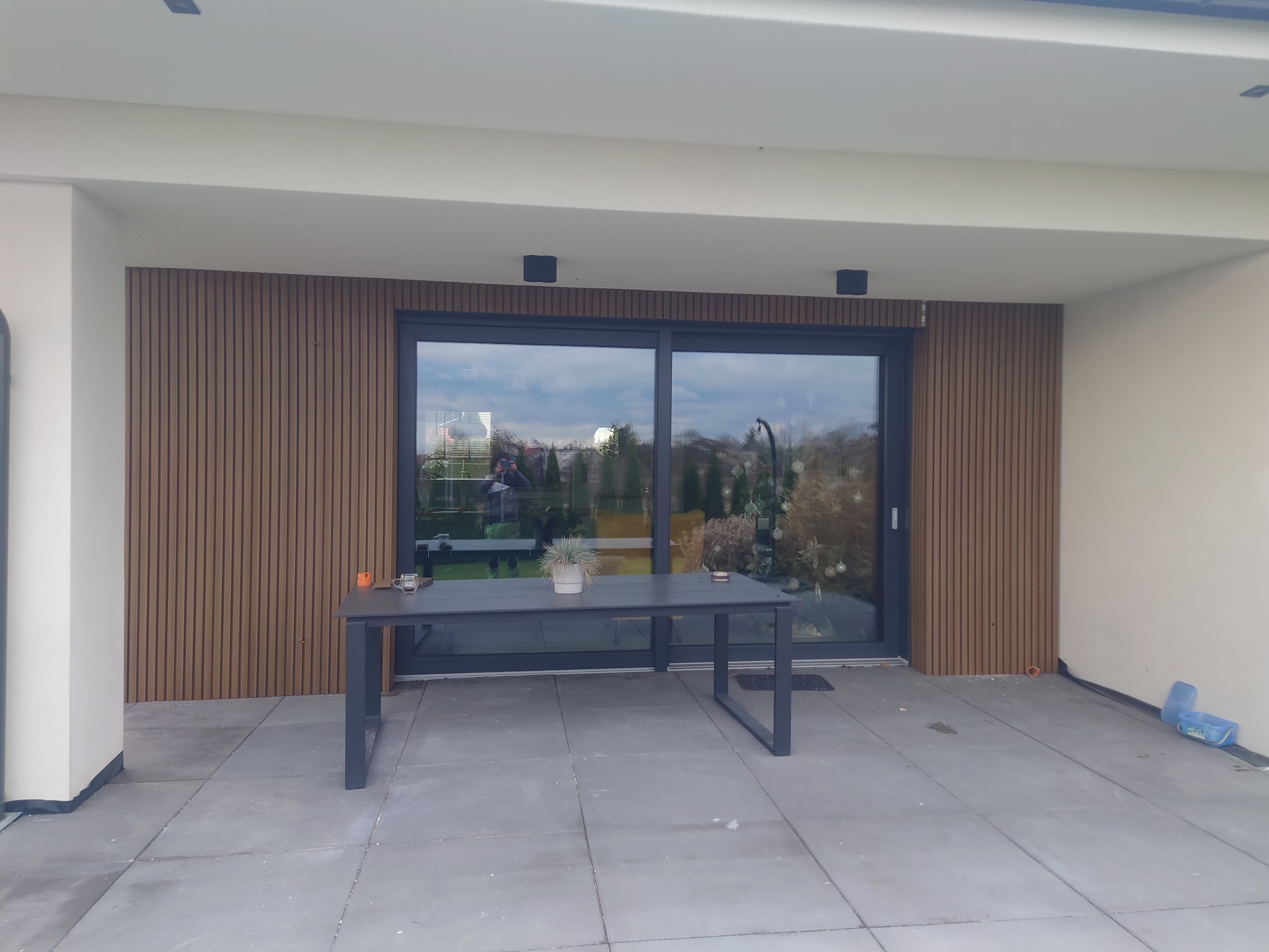 Nowoczesny taras z szarych płyt w Rogowo, z widocznym stołem ogrodowym i ścianą z drewnianymi panelami, przylegający do domu z dużymi przesuwnymi drzwiami.