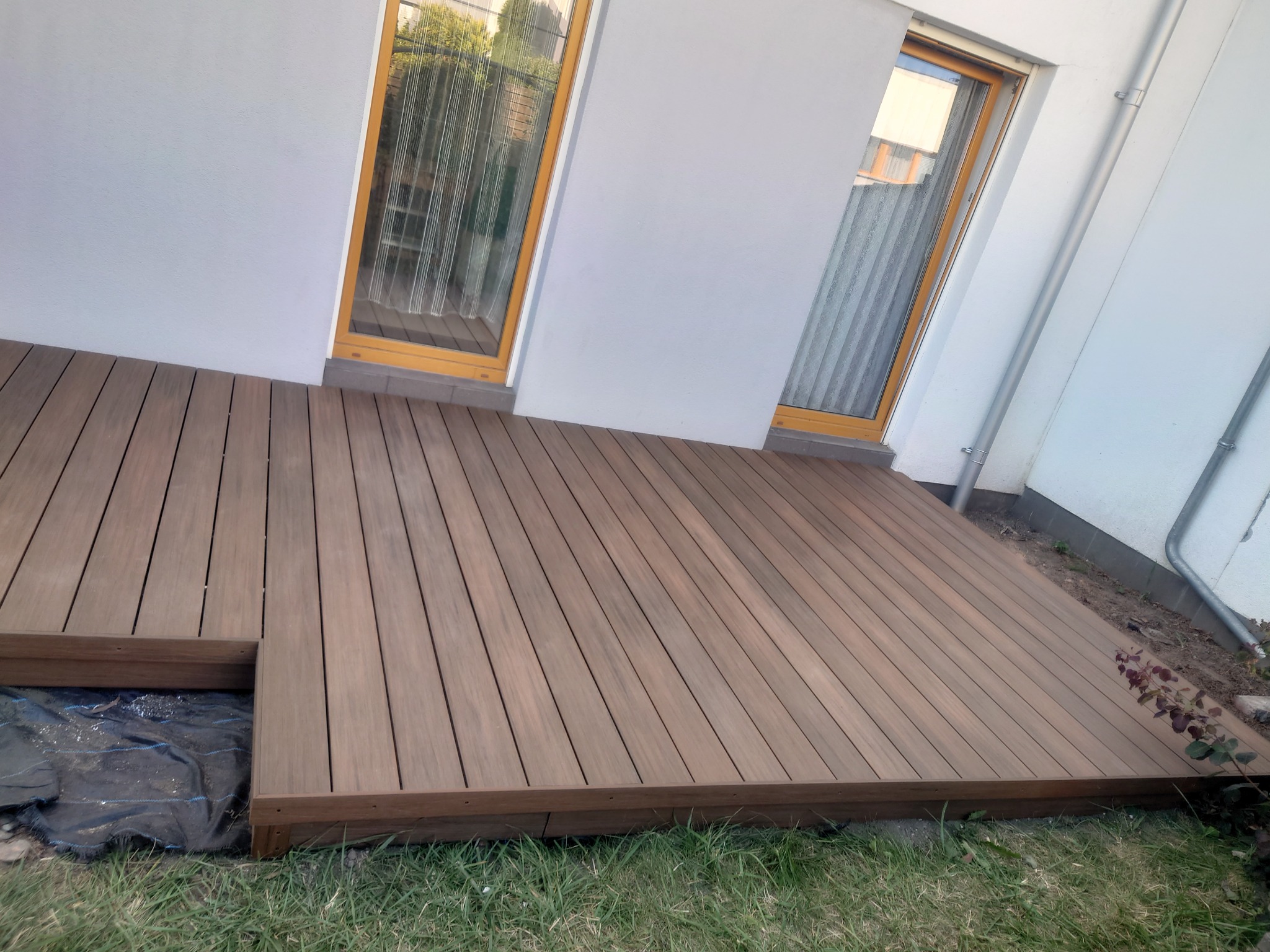 Nowoczesny taras z desek kompozytowych w odcieniach brązu, przylegający do białej ściany budynku z dwoma oknami, widoczne elementy trawnika i czarna folia pod konstrukcją.