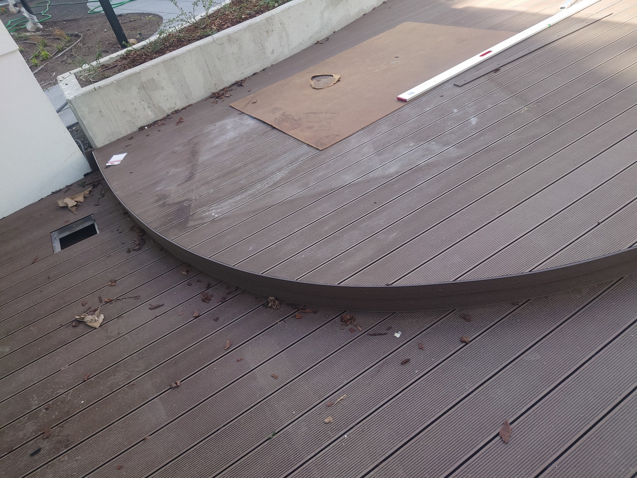 Półokrągły taras z desek kompozytowych w trakcie budowy, widoczna płyta z wyciętym otworem i poziomica, na deskach leżą liście
