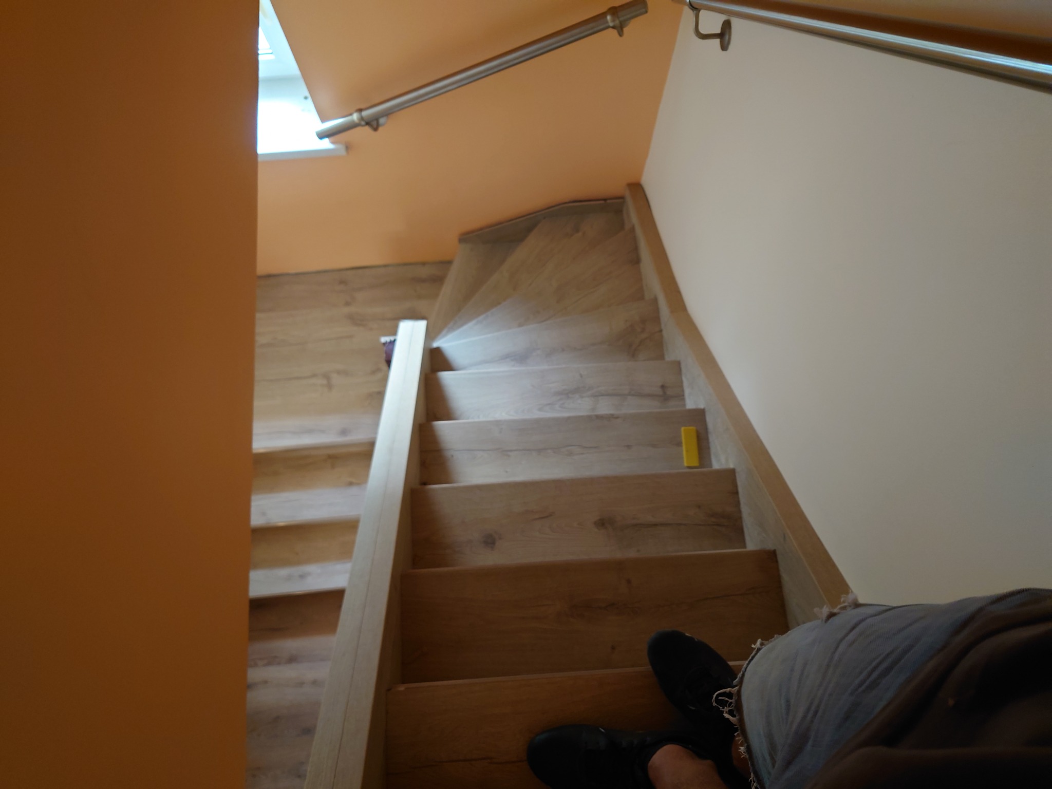 Widok z góry na drewniane schody zabiegowe z metalową poręczą, z fragmentem osoby w jeansach i czarnych butach na dolnym planie, oraz żółtym elementem na jednym ze stopni.