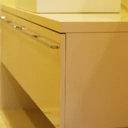 Beżowa szafka pod umywalkę z chromowanymi uchwytami na tle żółtych płytek z falistym wzorem, widoczna waga łazienkowa w tle.