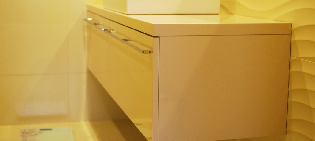 Beżowa szafka pod umywalkę z chromowanymi uchwytami na tle żółtych płytek z falistym wzorem, widoczna waga łazienkowa w tle.