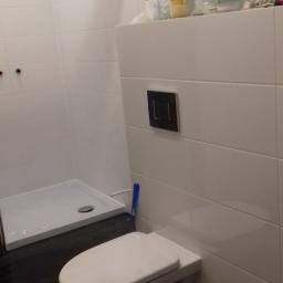 Krzysztof - Nowoczesna łazienka w trakcie wykańczania: podwieszana toaleta, białe płytki na ścianach, czarna podłoga i brodzik, widoczne materiały budowlane na górze.