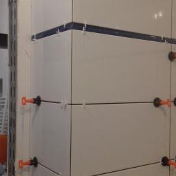 Krzysztof - Świeżo ułożone, białe, kwadratowe płytki ceramiczne na ścianie z użyciem krzyżyków dystansowych i poziomujących klipsów z pomarańczowymi klinami, widoczna listwa dekoracyjna.