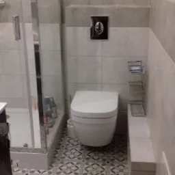 Wykończona łazienka z kabiną prysznicową, podwieszaną toaletą, mozaikową podłogą i szarymi płytkami ściennymi z ciemniejszym pasem dekoracyjnym.