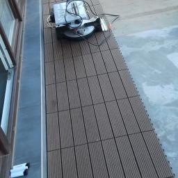 Krzysztof - Układanie paneli tarasowych z użyciem piły ukośnicy, obok leży czarny kot, widoczne otwarte okno balkonowe z pomarańczowym przedłużaczem.