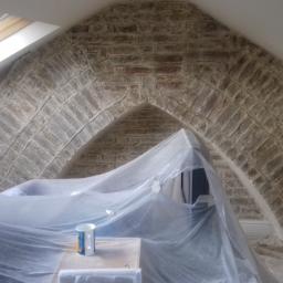 Krzysztof - Skośny strych z ceglanym łukiem w trakcie prac, zabezpieczony folią malarską, widoczny kubek na stole.