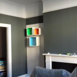 Krzysztof - Pokój po malowaniu ścian na szaro z białym pasem pod sufitem, zabezpieczone meble folią malarską, biały kominek, kolorowe półki na ścianie.