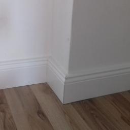 Krzysztof - Szczegół narożnika pokoju po remoncie: białe, profilowane listwy przypodłogowe na styku z panelami podłogowymi o wzorze drewna.