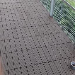 Krzysztof - Nowa podłoga z paneli kompozytowych na balkonie w Warszawie, widok z góry na ułożone panele i metalową barierkę, obok barierki widoczna zieleń, na podłodze leży rolka szarej taśmy klejącej.