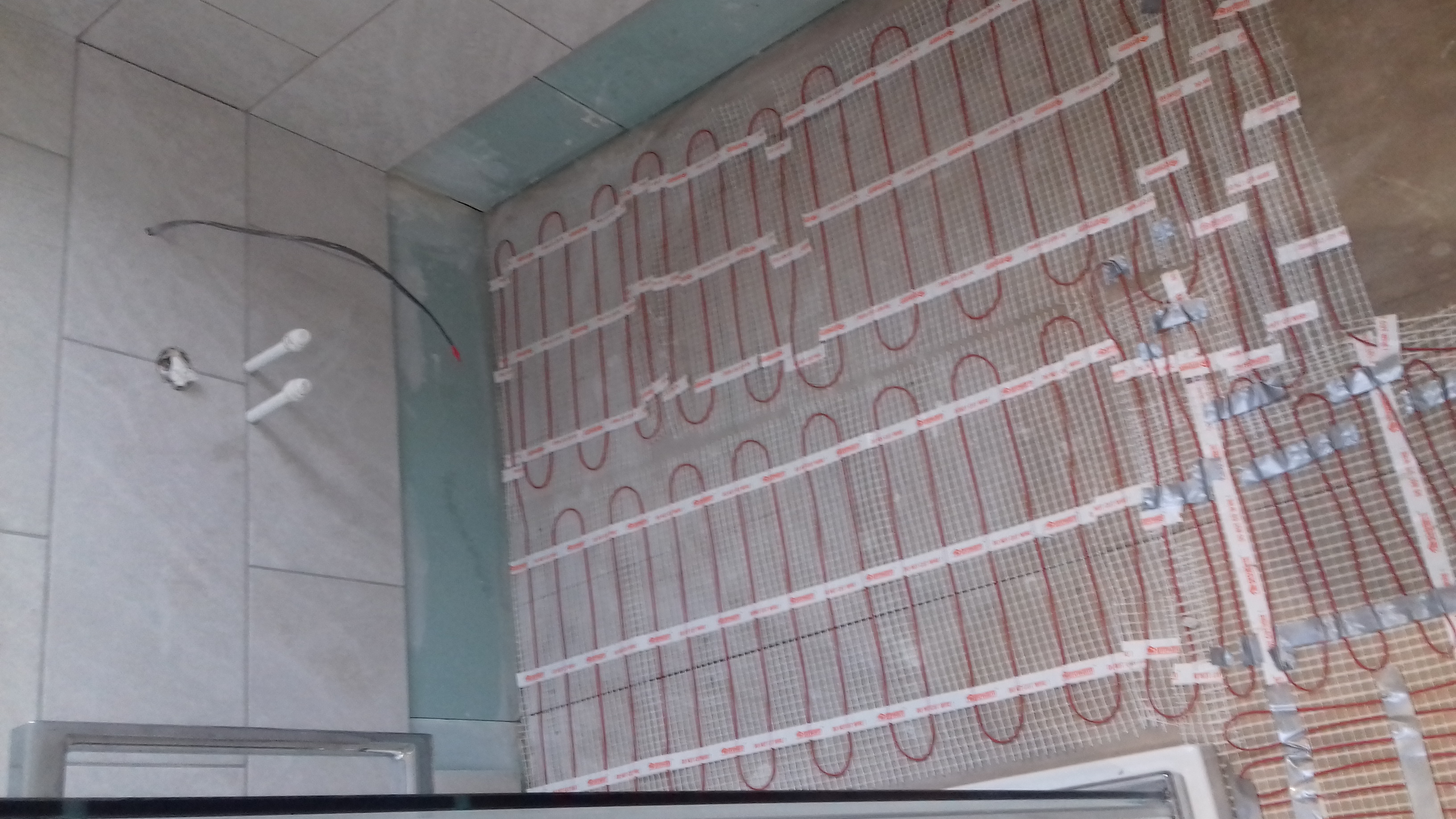 Przygotowanie do montażu ogrzewania podłogowego w łazience, widoczne kable elektryczne, siatka montażowa z taśmą mocującą oraz fragment ściany z ułożonymi płytkami.