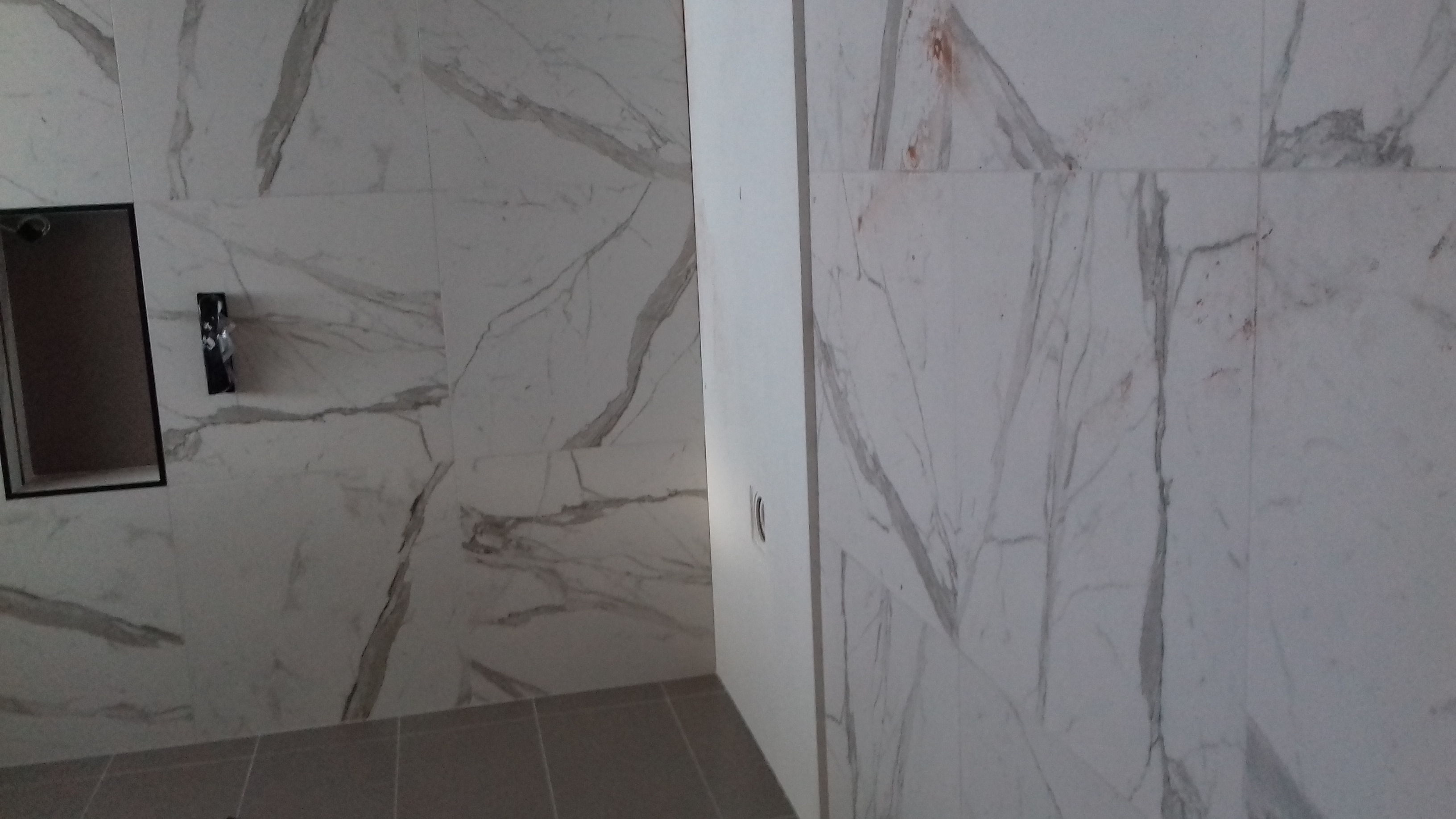 Ściana łazienki wyłożona białymi płytkami imitującymi marmur z szarymi żyłami; widoczna wnęka w ścianie z czarną ramką oraz fragment podłogi z szarych płytek.