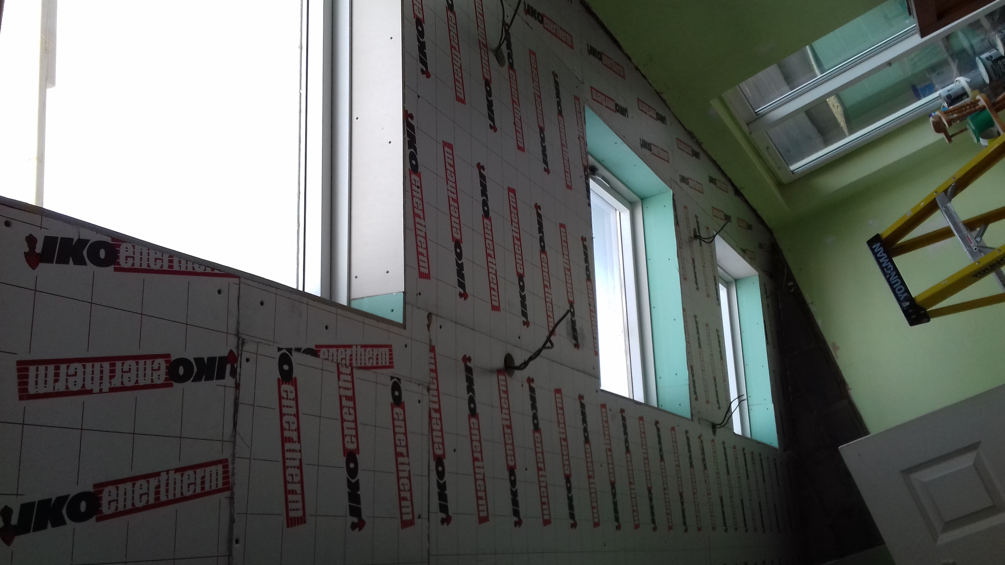 Ściana wewnętrzna z oknami w trakcie ocieplania, pokryta płytami izolacyjnymi z widocznym logo IKO enertherm, wystające przewody elektryczne, fragment drabiny i okna dachowego.