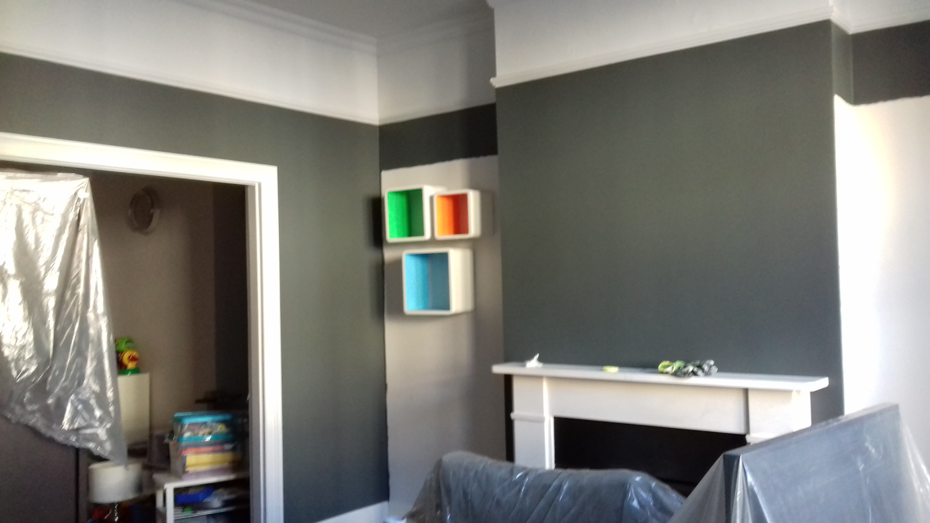 Pokój po malowaniu ścian na szaro z białym pasem pod sufitem, zabezpieczone meble folią malarską, biały kominek, kolorowe półki na ścianie.
