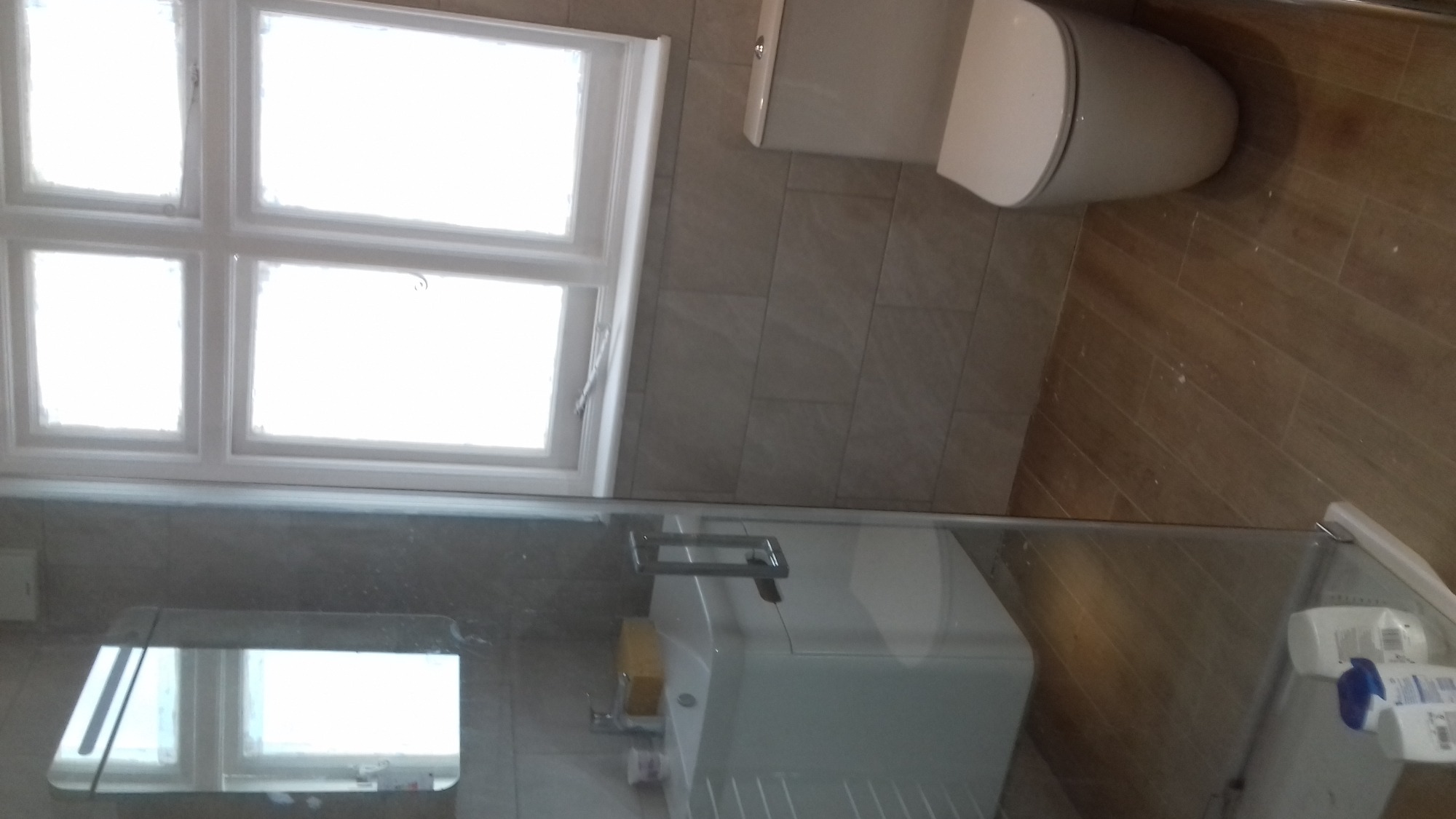 Wykończona łazienka z beżowymi płytkami, drewnopodobną podłogą, białą umywalką, toaletą i oknem z białymi ramami, widziana z góry przez szklaną kabinę prysznicową.