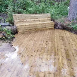 Nowa drewniana podłoga tarasowa z ławką w ogrodzie, pokryta wodą po deszczu, z widocznym fragmentem pnia drzewa i ogrodzeniem z gałęzi w tle.