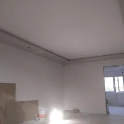 Pokój w trakcie wykańczania: ściany i sufit pokryte gładzią, widoczne płyty karton-gips na suficie z przygotowanymi otworami na oświetlenie, drabina oparta o ścianę, wstępne prace wykończeniowe.