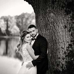 Fotografrawa - Czarno-białe zdjęcie pary młodej obejmującej się pod drzewem z widokiem na jezioro w tle, panna młoda w sukni z koronkowym tyłem, pan młody w smokingu z muchą.