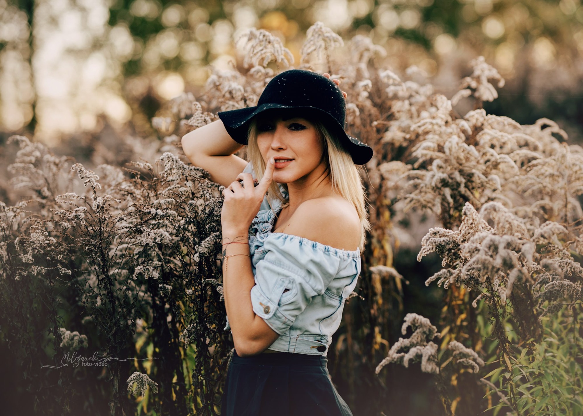 Portret kobiety w kapeluszu na tle kwitnących roślin, z palcem przy ustach i rozmarzonym spojrzeniem.