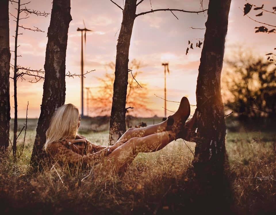 Kobieta z blond włosami opiera się o drzewo na tle zachodzącego słońca i turbin wiatrowych, ubrana w brązową kurtkę i buty.