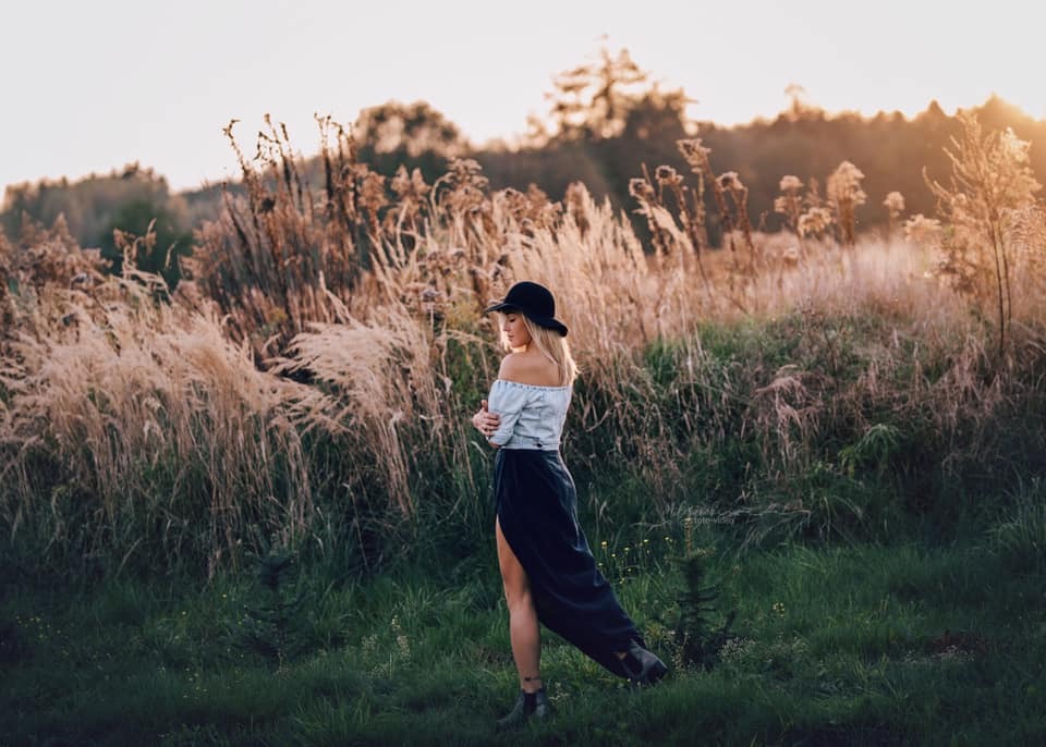 Kobieta w kapeluszu, bluzce z odkrytymi ramionami i długiej spódnicy z rozcięciem, pozuje na tle wysokich traw w promieniach zachodzącego słońca.