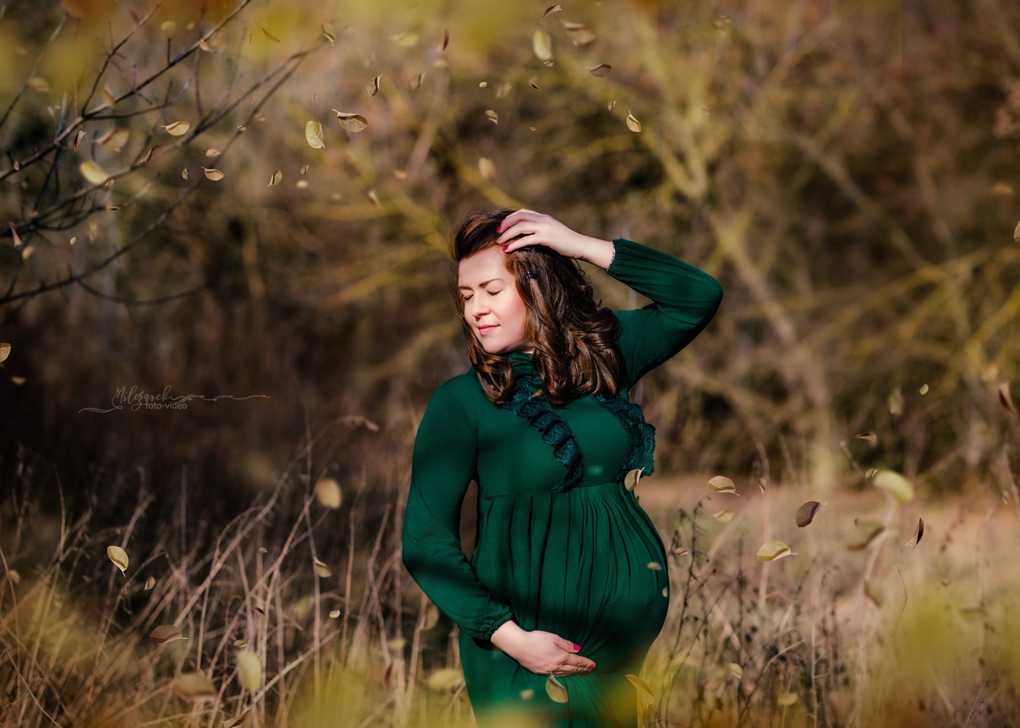 Kobieta w zielonej sukience ciążowej pozuje na tle jesiennego krajobrazu w okolicach Rawy Mazowieckiej, opadające liście tworzą efektowną aurę.