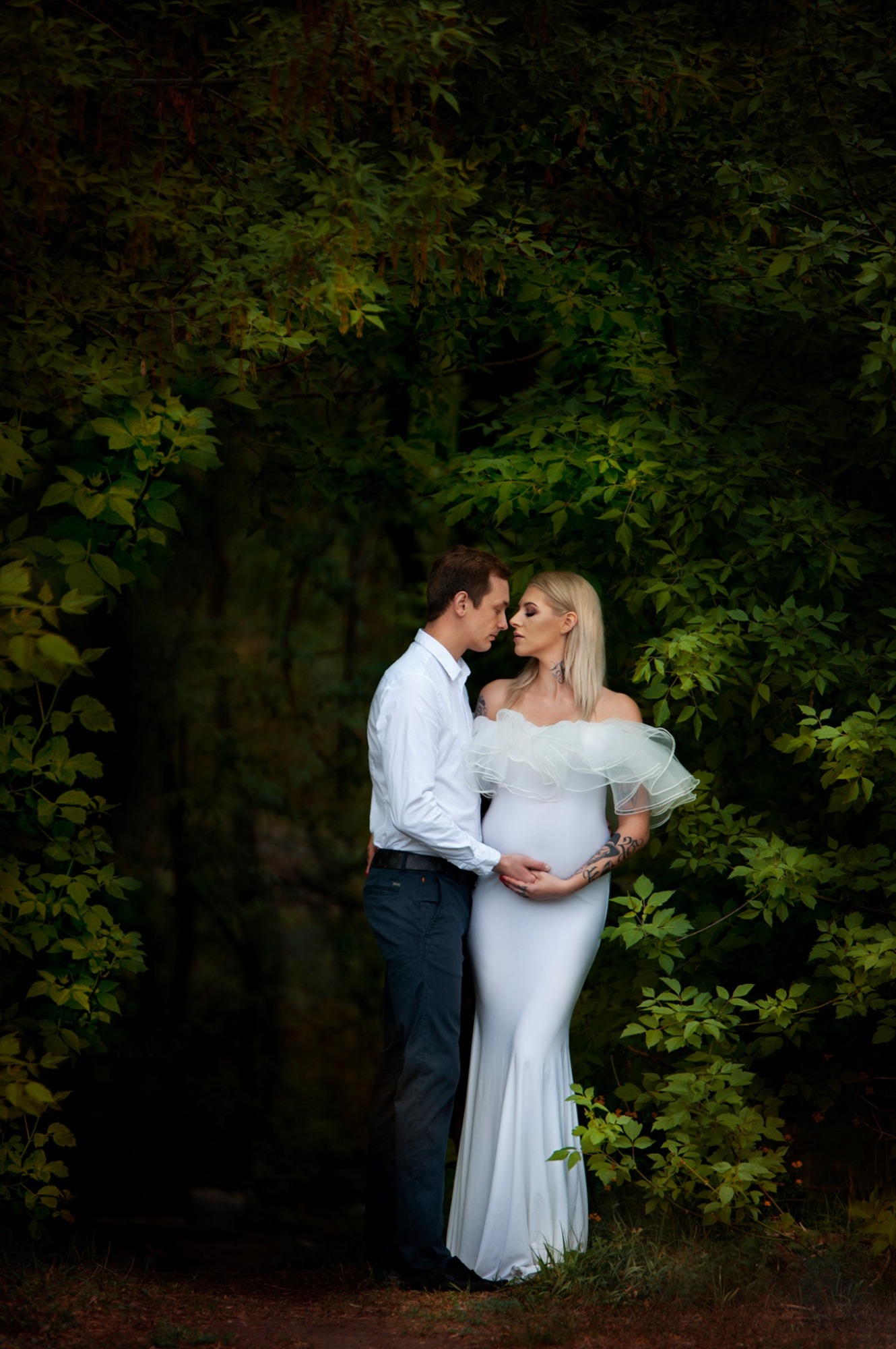 Przyszła mama w białej sukni z falbanami i przyszły tata obejmujący brzuch na tle zieleni