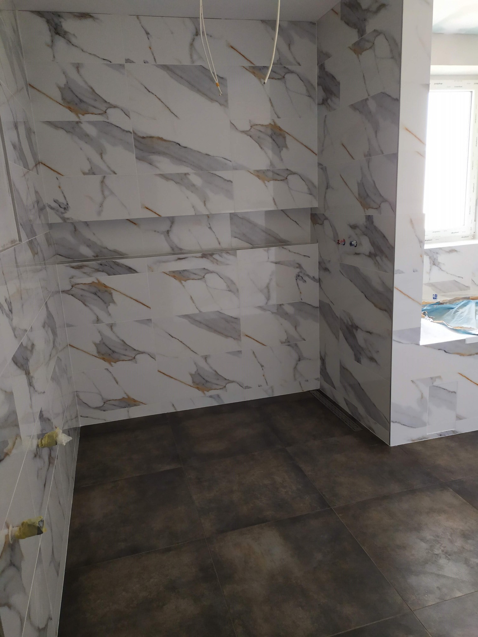 Wykończona łazienka w Kolbuszowej z białymi płytkami imitującymi marmur na ścianach, z wnękami i ciemnobrązowymi płytkami na podłodze, widoczne zabezpieczone taśmą punkty instalacyjne.
