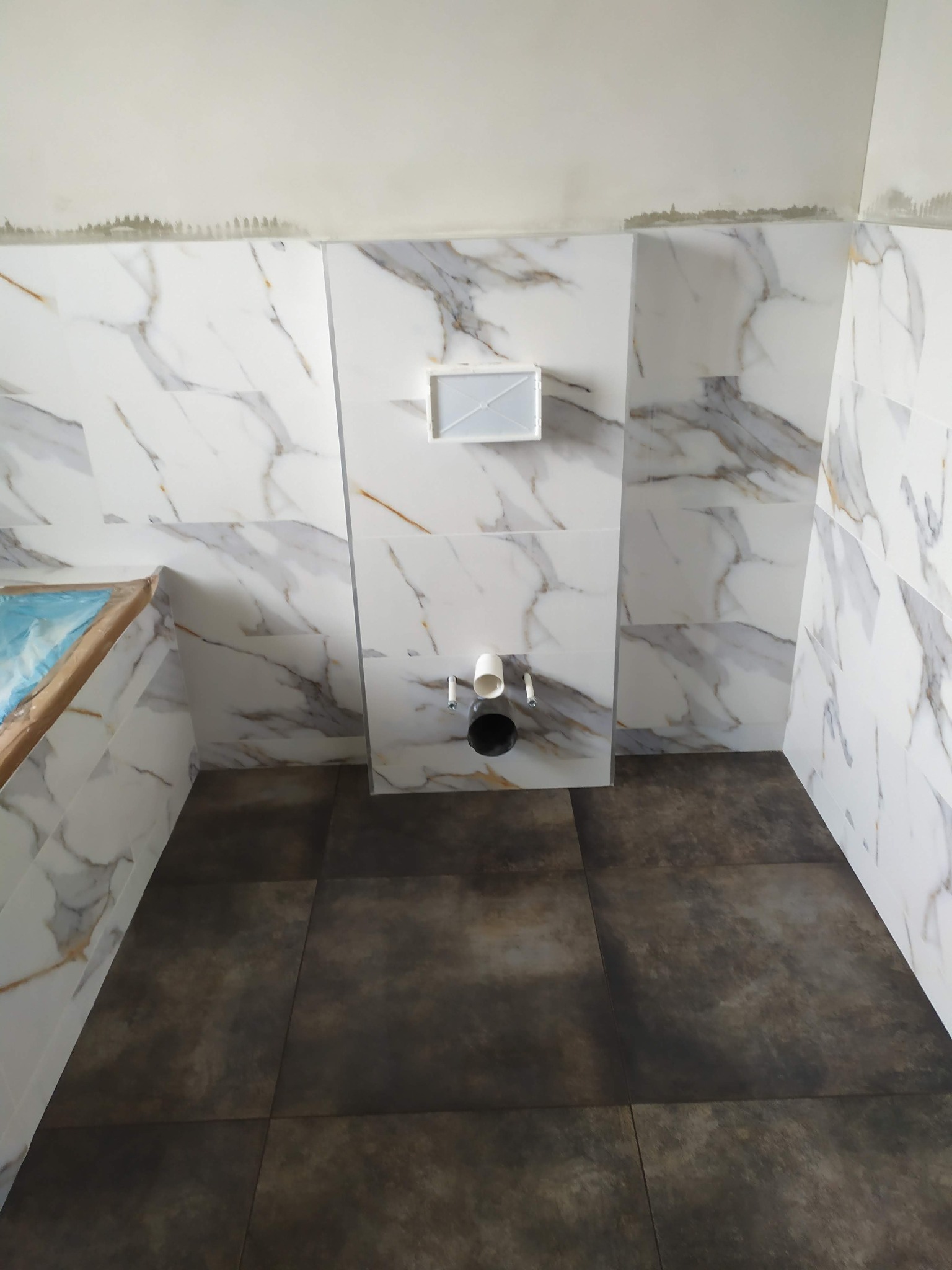 Wnętrze łazienki w trakcie prac wykończeniowych, widoczne białe płytki imitujące marmur na ścianach, ciemne płytki na podłodze oraz częściowo zamontowana toaleta podtynkowa z widocznymi rurami.