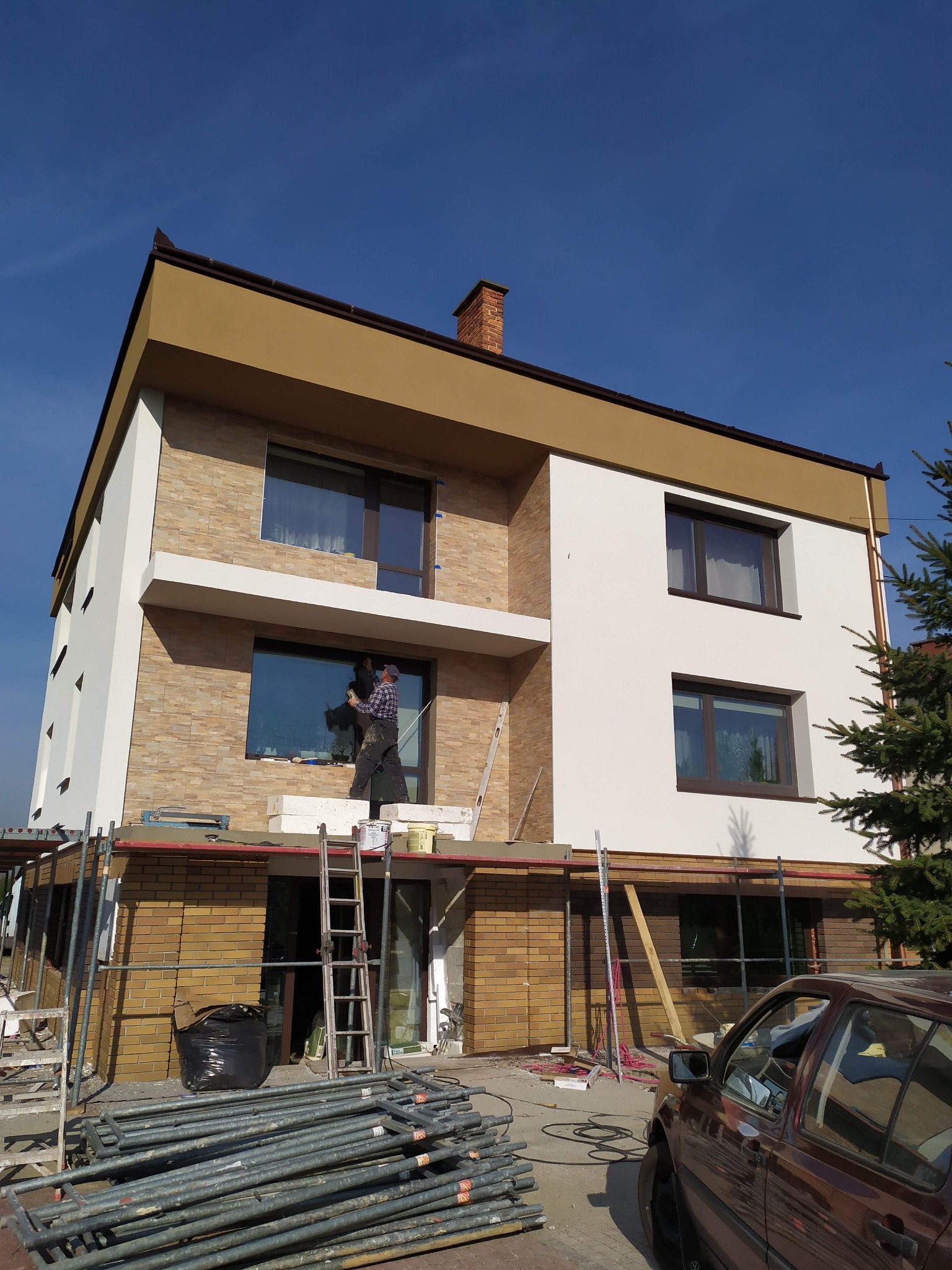 Dwukondygnacyjny dom w trakcie ocieplania elewacji, widoczny pracownik na rusztowaniu przy oknie, fragmenty elewacji z jasnego kamienia i białego tynku, w tle błękitne niebo.