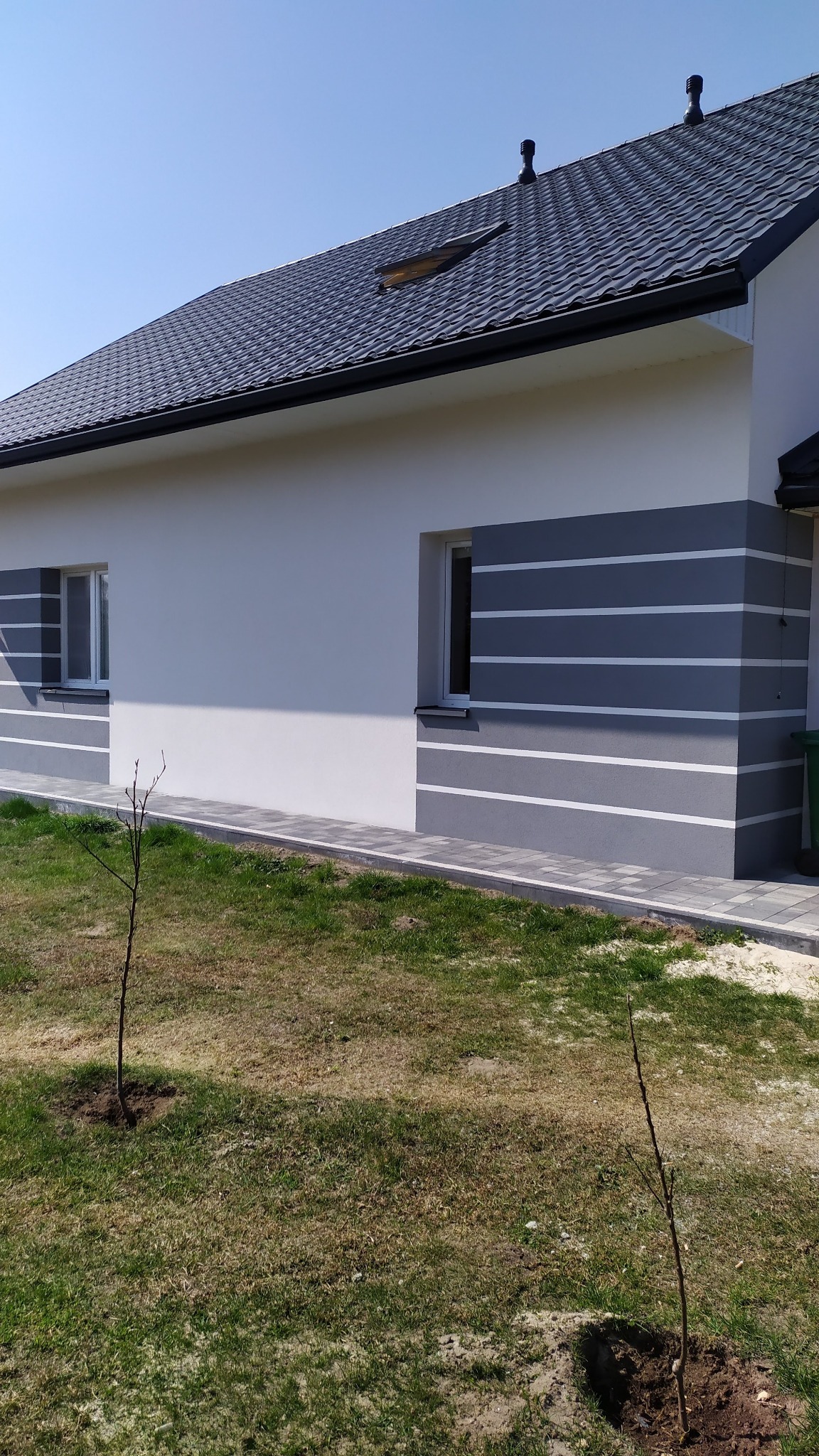 Elewacja budynku jednorodzinnego z szarymi pasami, świeżo posadzone drzewka na trawniku.