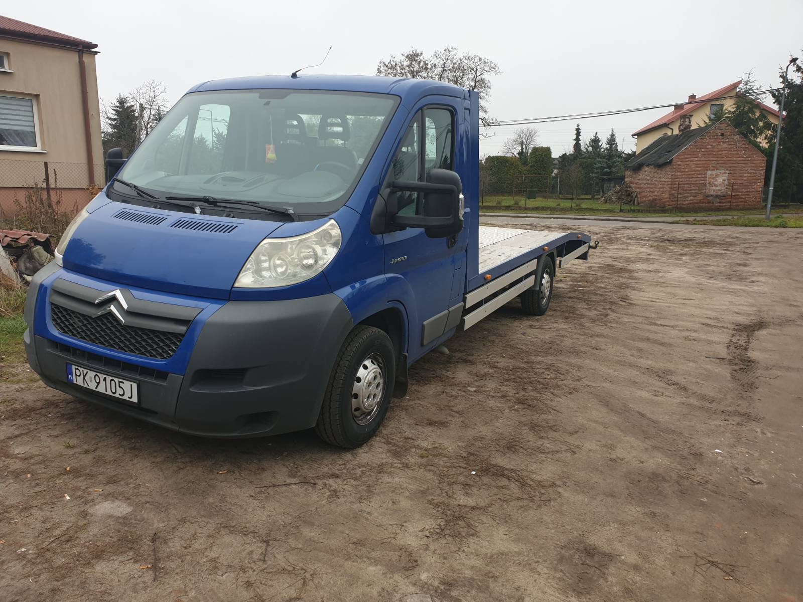 Niebieski samochód dostawczy z platformą do transportu pojazdów, zaparkowany na placu z ubitej ziemi, widok z przodu na tle budynków.