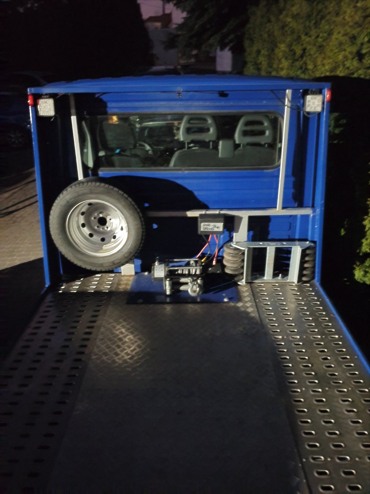 Niebieska laweta z widoczną platformą, kołem zapasowym i wyciągarką, oświetlona światłami LED, sfotografowana nocą.
